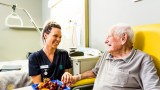 An elderly man sits in a yellow chair at a table with a bowl of fruit, holding the hand of a young female Mercy Health worker. The man and woman are smiling at each other.