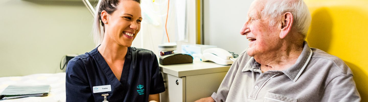 An elderly man sits in a yellow chair at a table with a bowl of fruit, holding the hand of a young female Mercy Health worker. The man and woman are smiling at each other.