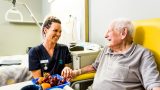 An elderly man sits in a yellow chair at a table with a bowl of fruit, holding the hand of a young female Mercy Health worker. The man and woman are smiling at each other.