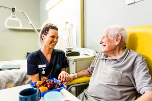 An elderly man sits in a yellow chair at a table with a bowl of fruit, holding the hand of a young female Mercy Health worker. The man and woman are smiling at each other.