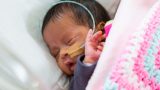 Close up of a new born baby attached to a tube from the nose asleep tucked into pink blankets.