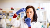 A medium close up of a woman in a lab wearing blue gloves and white lab coat holding a test tube.