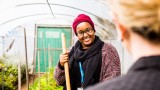 An over the shoulder photo of a female Mercy Health worker in a green house holding a gardening tool and smiling at a colleague