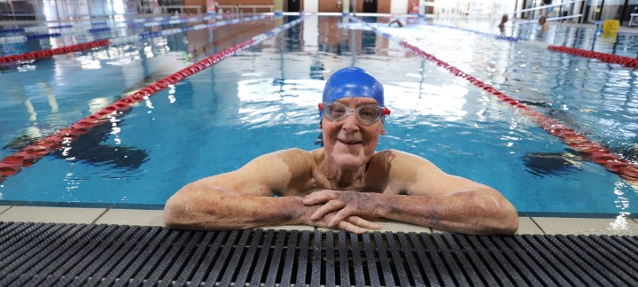 95-year-old Mercy Health Home Care client Father Chris Baker, enjoying swimming in a pool with his arms leaning up whilst smiling at camera.
